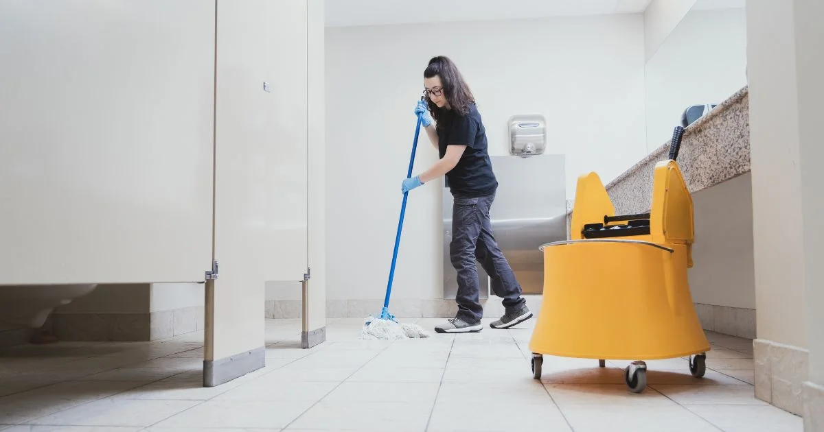 Worker wet mopping a restroom floor with bucket nearby